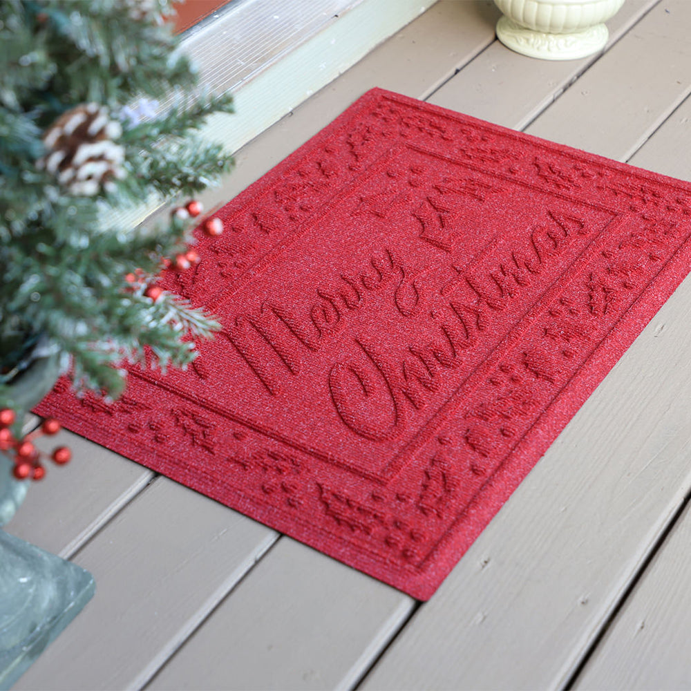 Red 'Merry Christmas' WaterHog doormat on a wooden floor with a Christmas tree for festivities