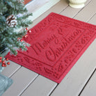 Red 'Merry Christmas' WaterHog doormat on a wooden floor with a Christmas tree for festivities