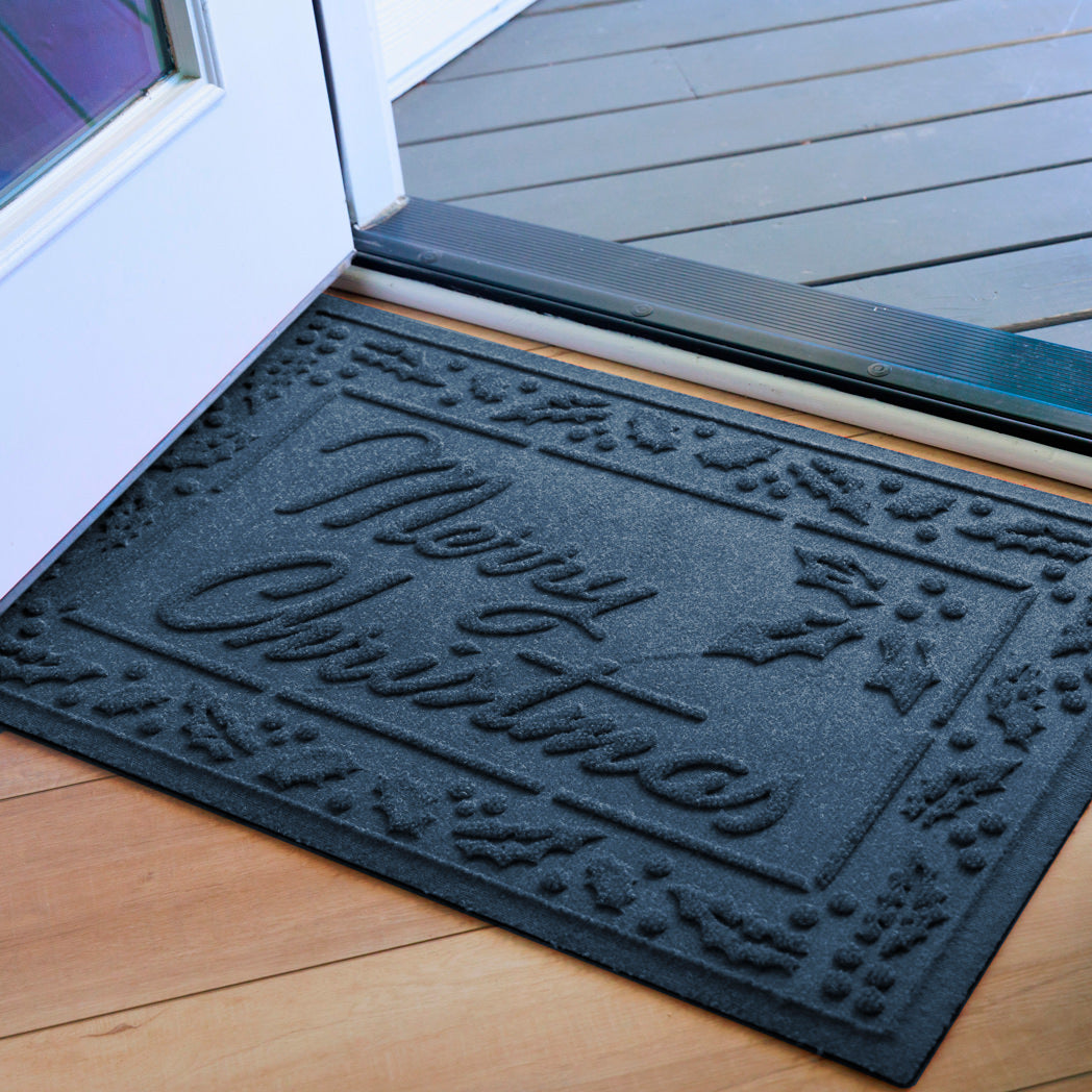 Deep blue doormat with  Merry Christmas' text surrounded by a Holly border on a wooden floor inside open front door. .