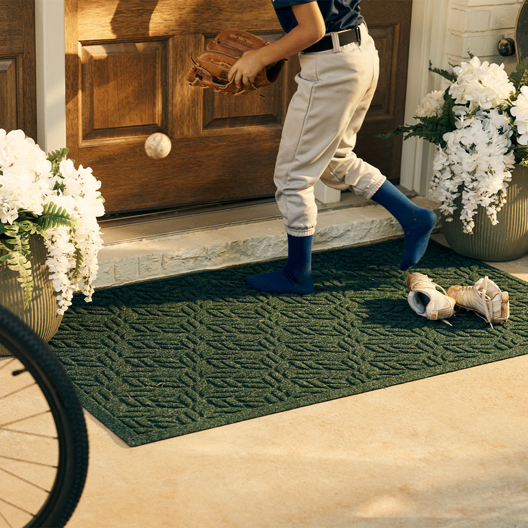 Young boy in baseball uniform with glove and ball, stepping out of his dirty cleats on a evergreen WaterHog canopy doormat.