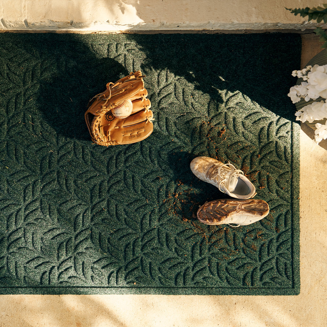 Quality WaterHog Canopy doormat in deep green, with a baseball glove, ball, and dirty cleats laying on mat.