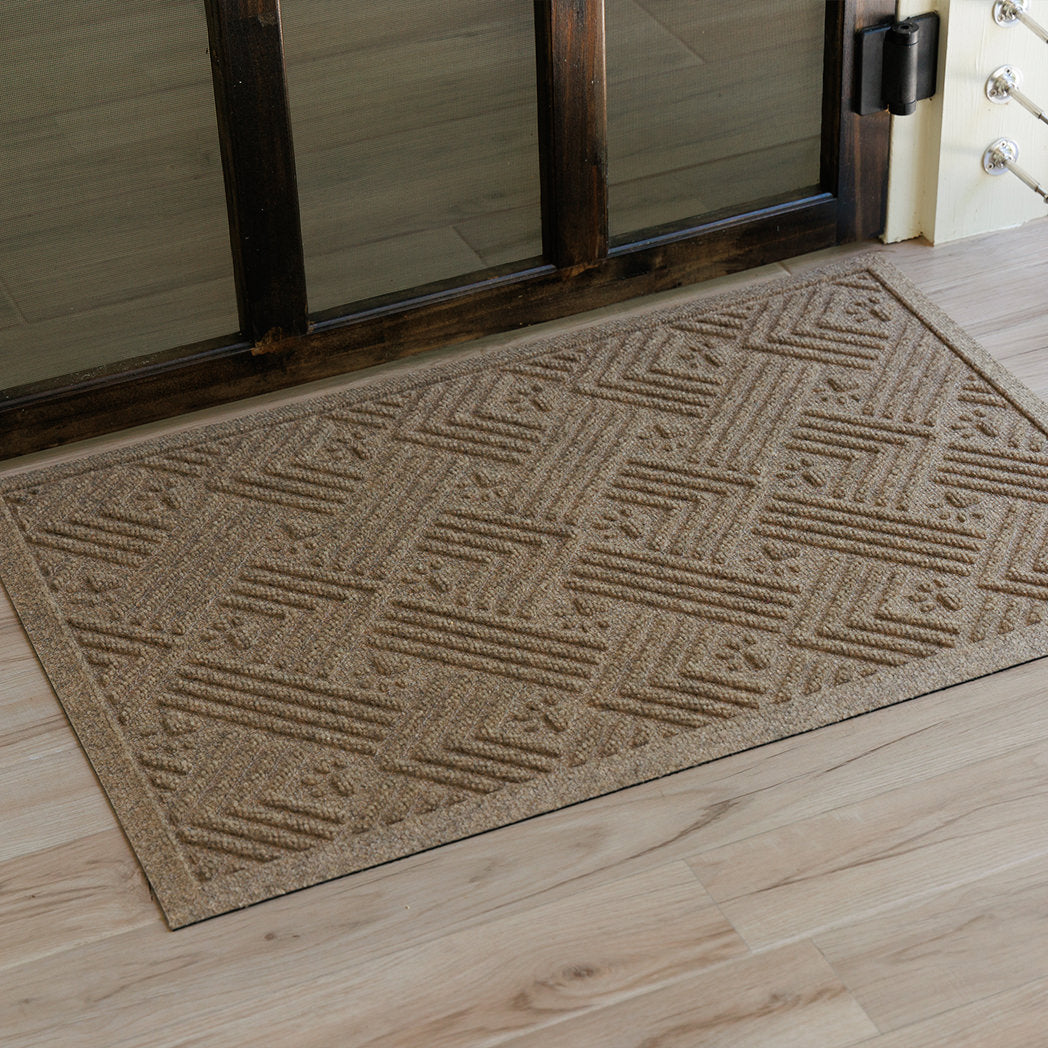 Overhead view of a tan WaterHog Cat Litter Mat placed just inside a dark wood-framed doorway on light wood flooring. The mat’s textured surface features a repeating pattern of angled ridges and small paw prints.