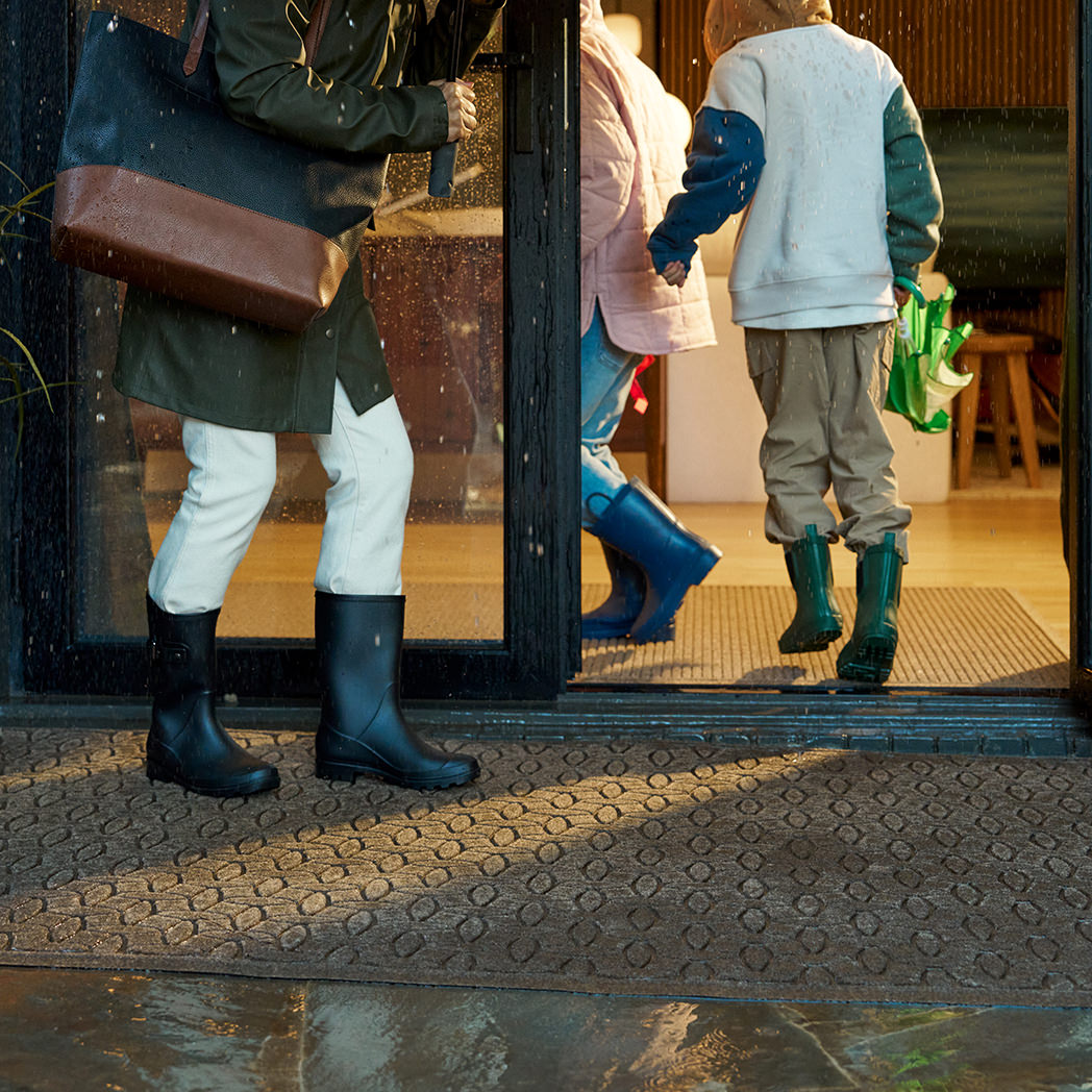 Large WaterHog Doormat in front of sliding glass doors. Rain is falling and three people in rubber boots are walking inside across the mat.