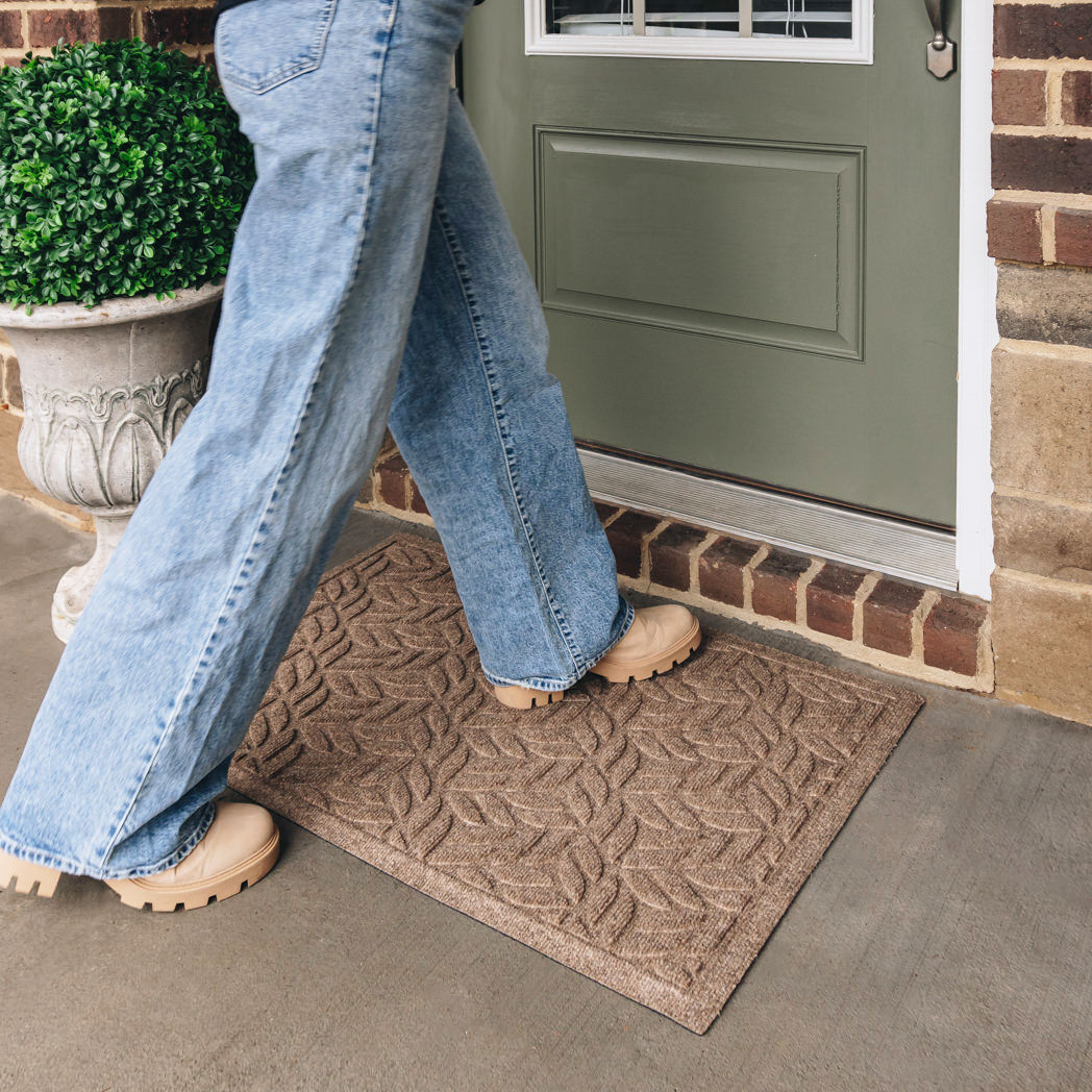 Front door with an OG WaterHog medium indoor/outdoor doormat in a light tan leaf petal design placed in front and a person in jeans and boots walking across.