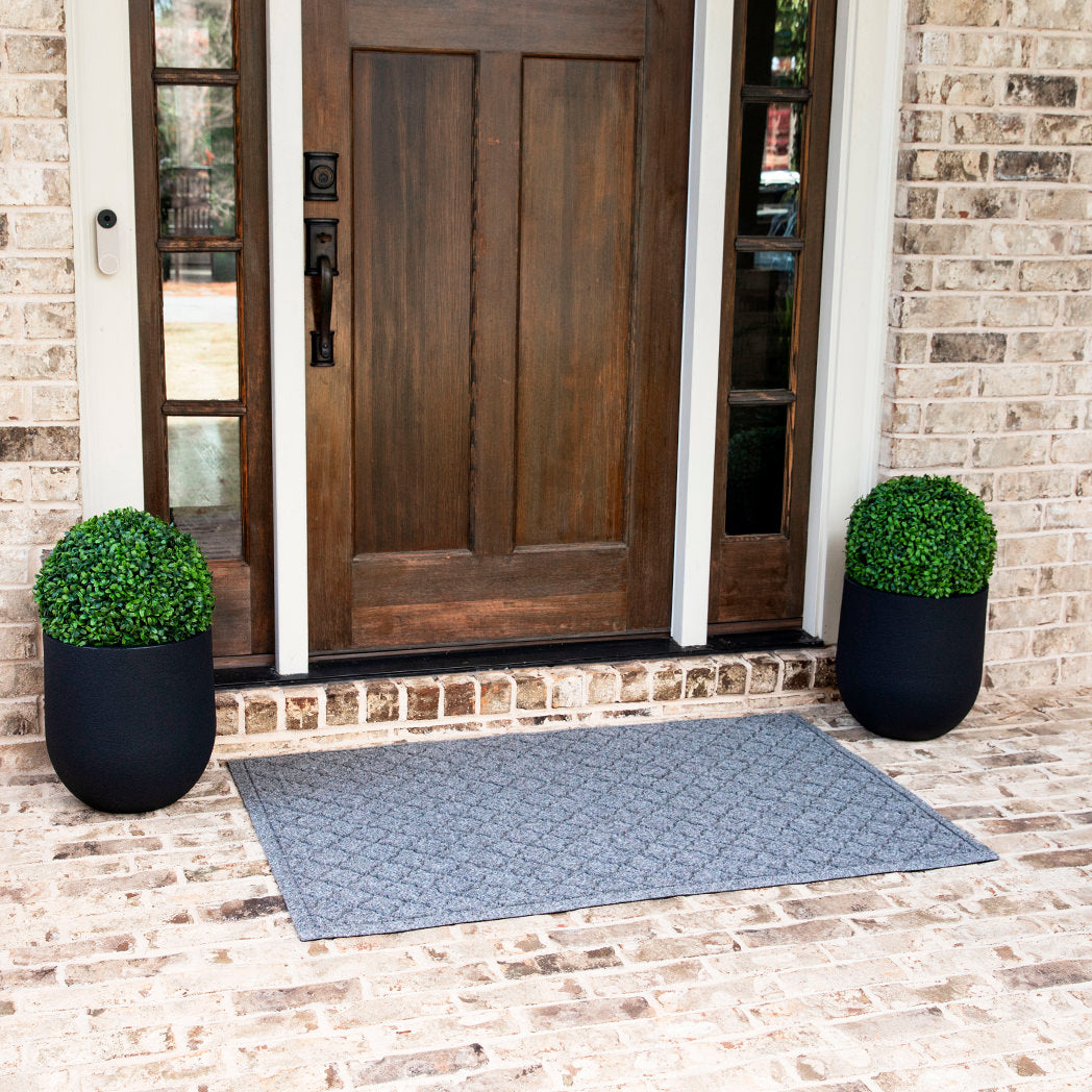 Beautiful deep brown front door with a large eco-friendly all-weather WaterHog doormat in medium grey with vibrant green plants on either side.