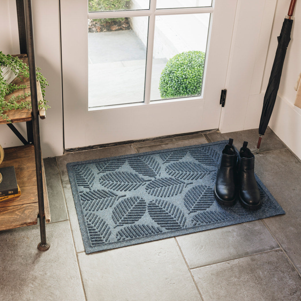 A medium-sized durable WaterHog doormat is placed inside in front of a single door that leads outside. The navy  blue doormat with a leafy design has a pair of black boots on top.