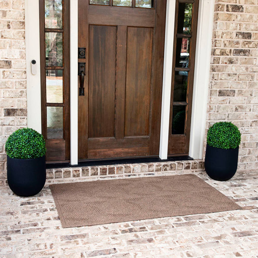 A large-sized, tan-colored WaterHog door mat in a leafy design, placed on brick flooring, outside a single door with sidelights.