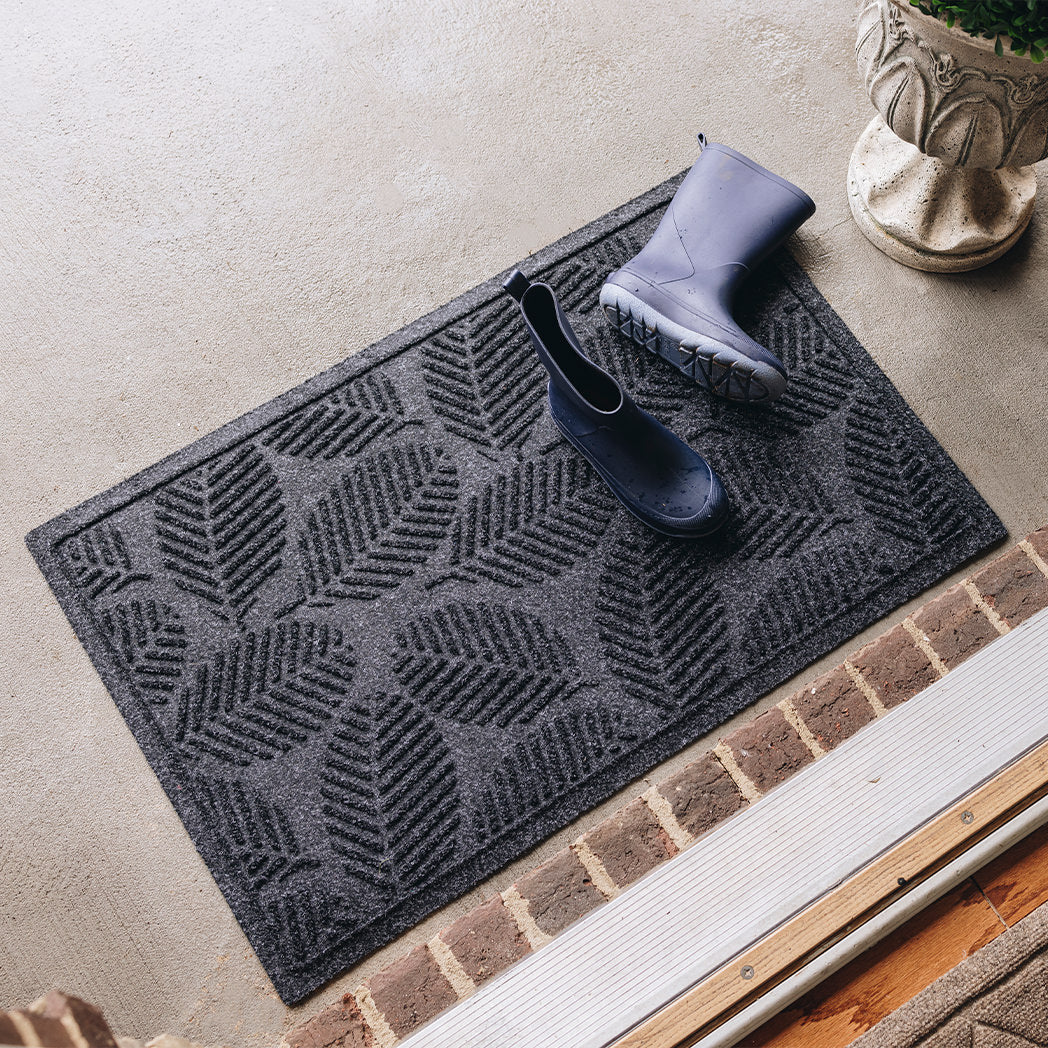 A medium-sized, durable WaterHog doormat in a dark gray/black color with a leafy design, placed outside on concrete flooring with a pair of navy blue boots with a few water spots on them.