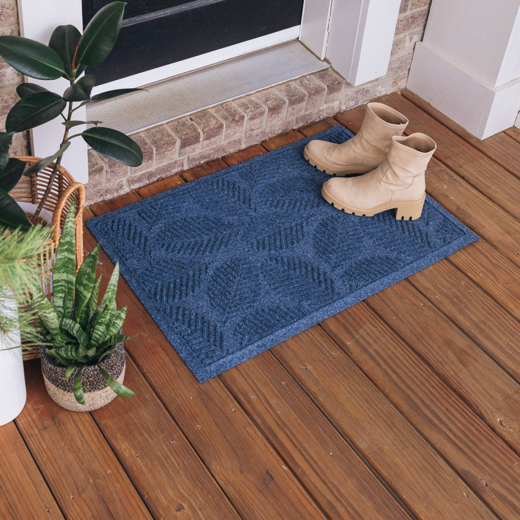 A medium-sized, all-weather WaterHog doormat in a navy blue color with a leafy design. The doormat is placed outside in front of a single door with a pair of boots placed on top of it.