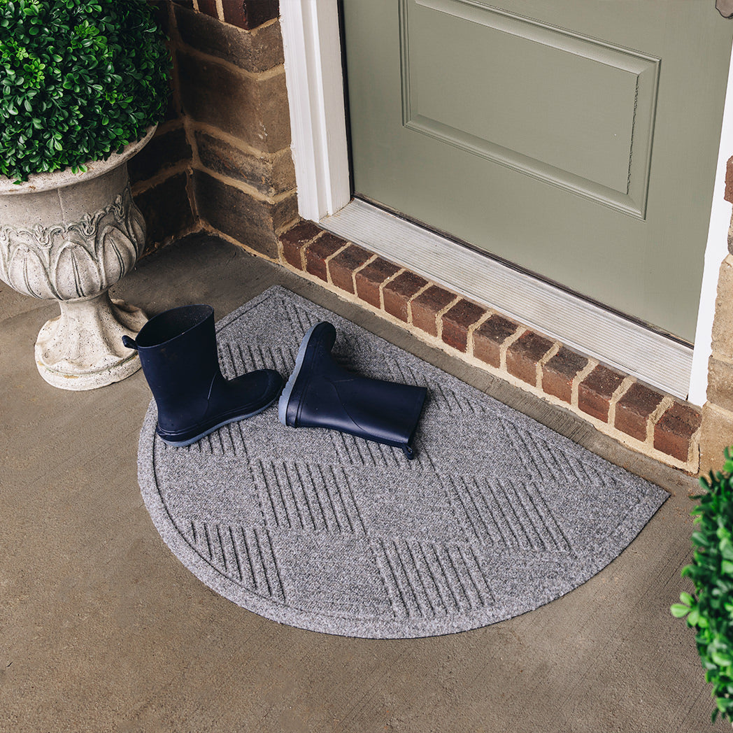 A medium grey half-round doormat with a linear crisscrossing design is placed on concrete flooring outside a single door with a pair of navy blue rainboots sitting on top.