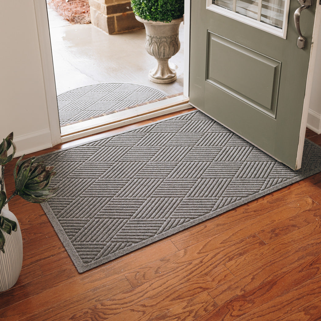 A large-sized, medium grey WaterHog doormat with a crisscrossing, linear design placed inside a single door on wood flooring, with the door open.