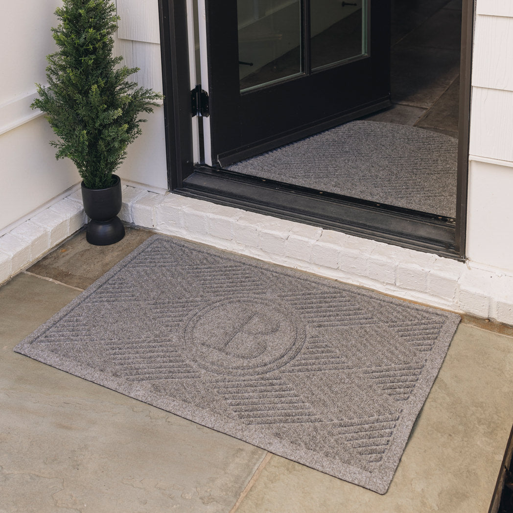 A medium grey monogrammed WaterHog doormat, with a diamond design, is placed on tile flooring outside a single door; the door is open to show a half-round doormat of the same color and pattern on the inside of the door.