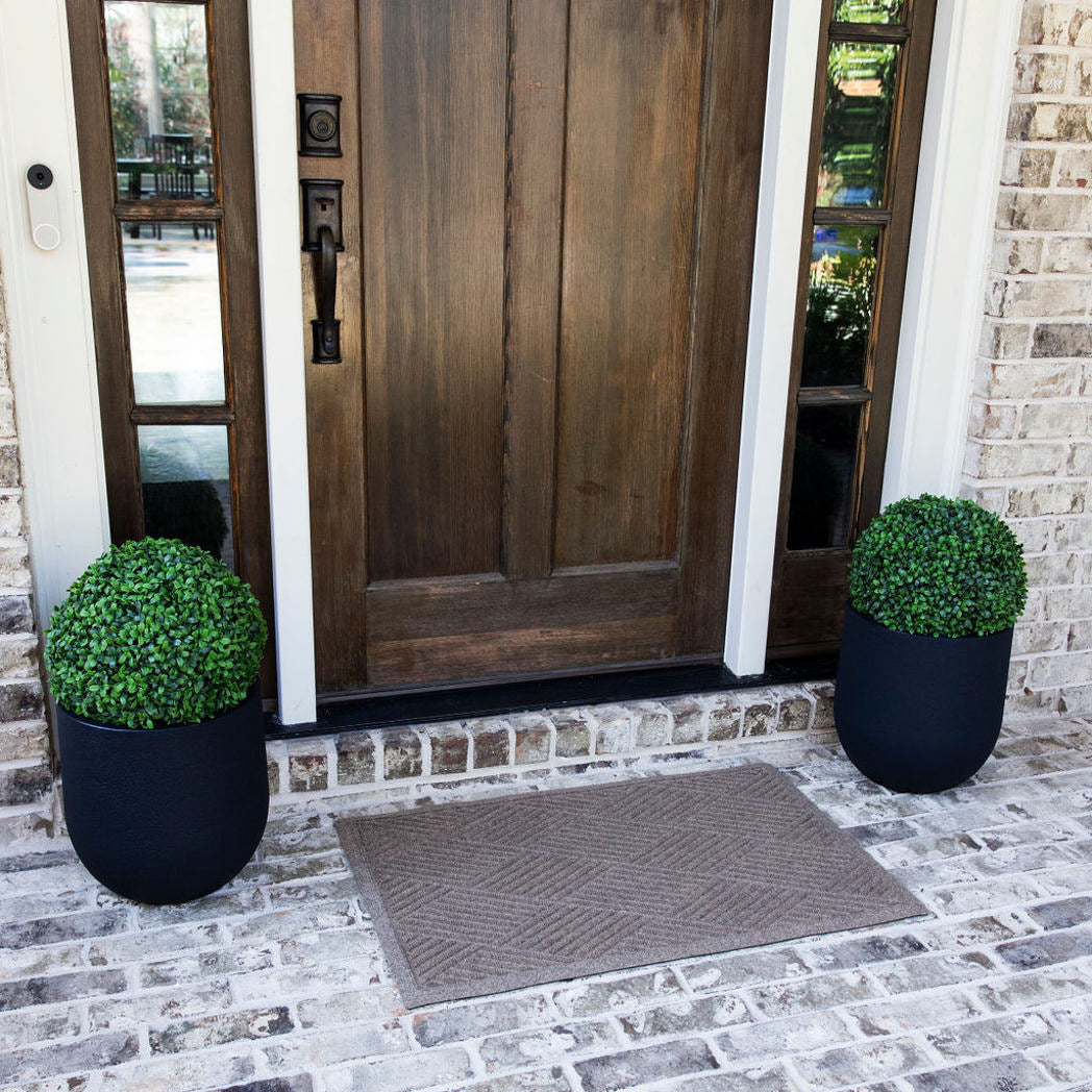 A medium-sized OG WaterHog door mat in medium grey with a crisscross, linear design sitting on brick flooring outside a single door with sidelights.