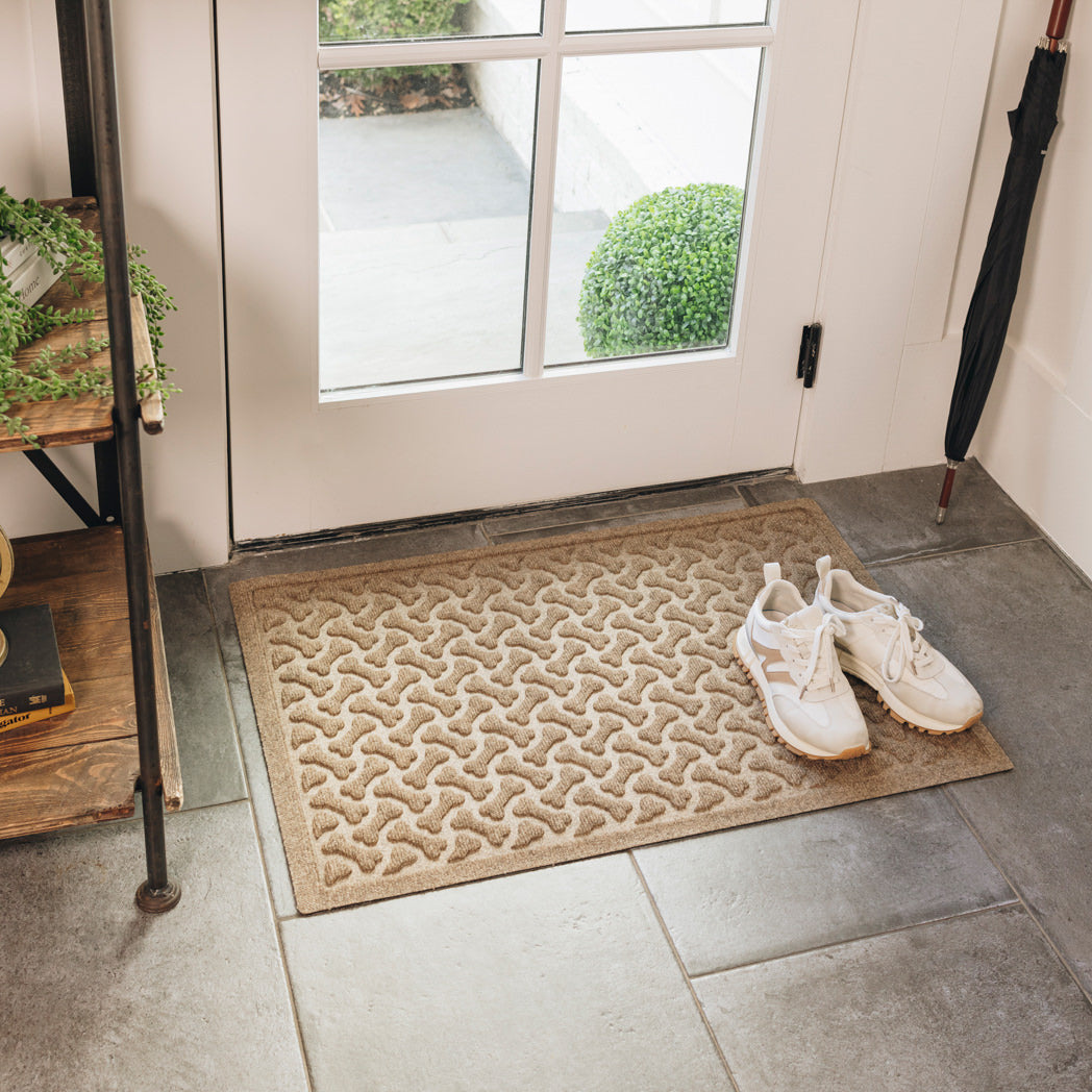 A light golden, medium-sized durable WaterHog mat, with a repeating dog bone pattern, is placed on tile flooring inside a single door with a pair of white sneakers sitting on it.