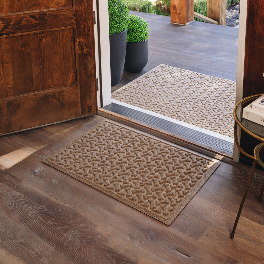A light tan all-weather doormat, with a repeating dog bones pattern , is placed on wood flooring inside a single door; the door is open to show a 3x5 outdoor doormat of the same color and pattern on the outside of the door.