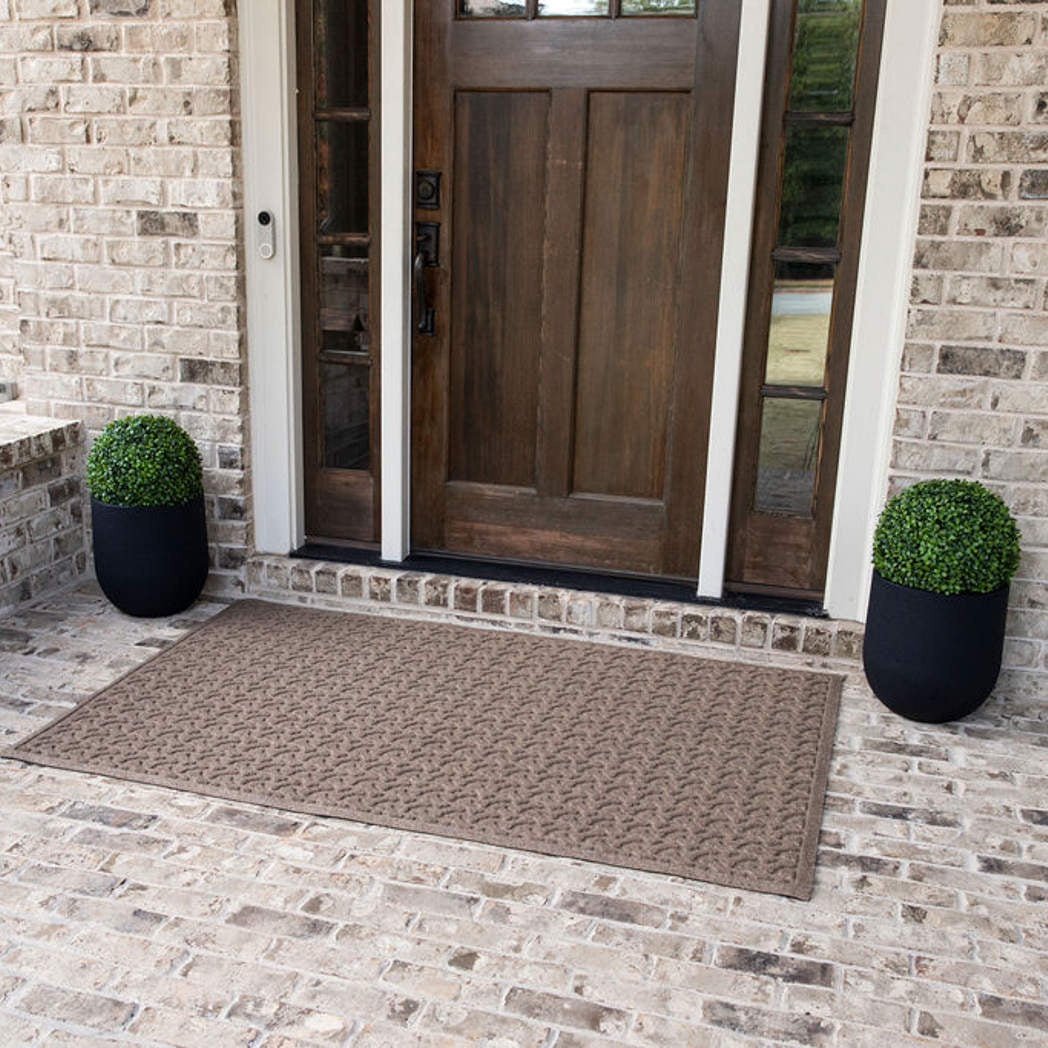 An extra-large outdoor mat in a light tan color with a repeating dog bone pattern placed on bricking flooring outside a single door with sidelights.