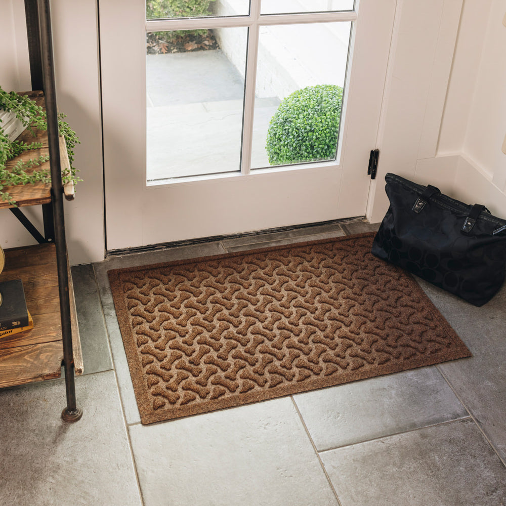 A medium-sized, brown WaterHog doormat, with a repeating dog bone pattern, is placed on tile flooring inside a single door with a black bag sitting off to the side.