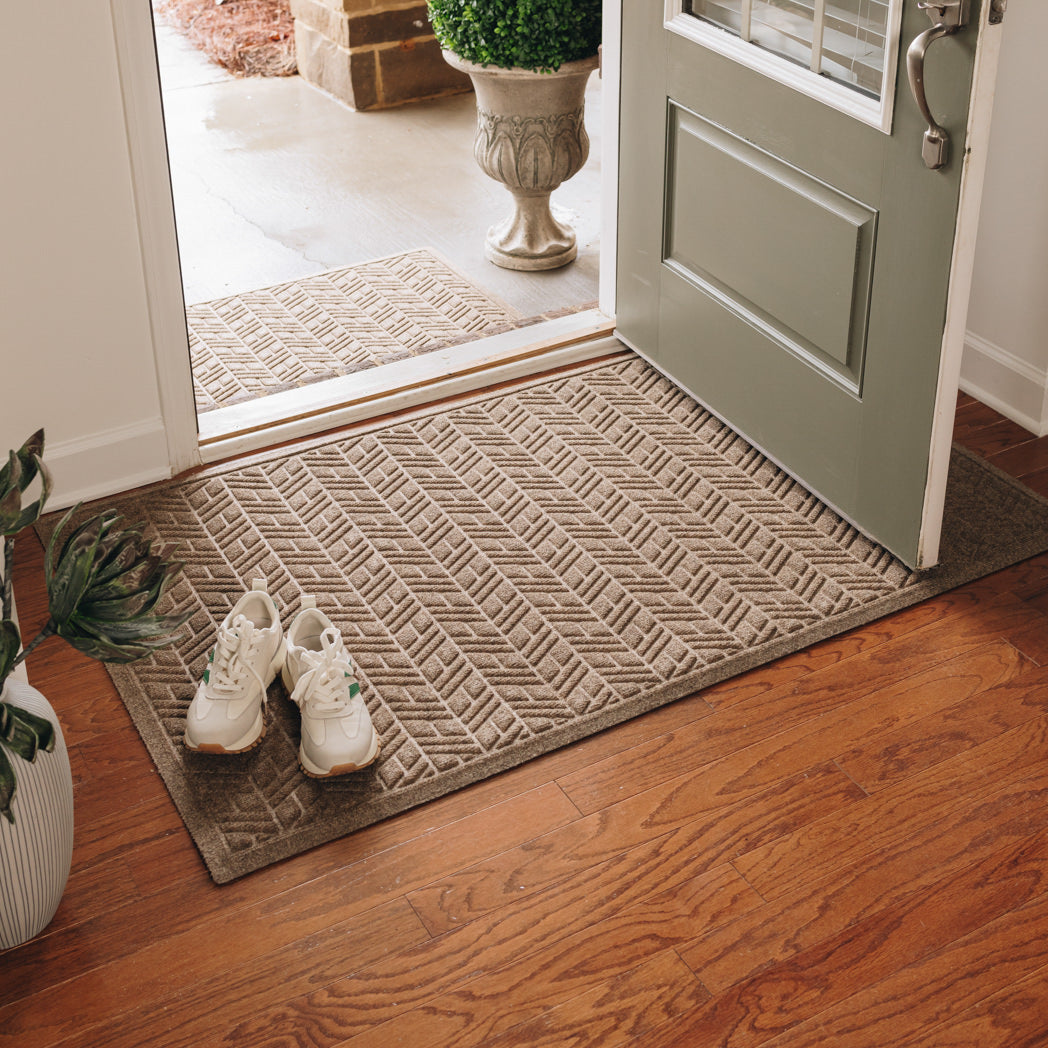 A brown WaterHog High Rise extra-large doormat with an elevated chevron-like design displayed on wood flooring inside an entrance way, and a medium 2 by 3 version displayed outside on concrete flooring.
