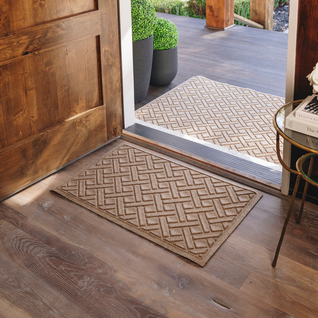WaterHog Lattice 2x3 indoor/outdoor mat in tan, featuring a crisscross geometric design, displayed on wood flooring in front of a single door inside, with a large version outside.