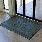 Overhead view of a blue-grey WaterHog Ship’s Anchor doormat laid just inside a black-framed glass door on light wood flooring. The photo shows the mat’s raised anchor emblem and braided border, evenly lit by soft daylight from the doorway.