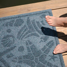 Close up of WaterHog's Beachcomber mat in blue with a seashell design on a deck with young girls feet in frame