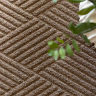 A close-up of the WaterHog Diamonds Doormat in a camel tan color shows its raised, angled-line pattern forming repeating diamond shapes. Each ridge has a corded texture that catches light and shadow, giving the mat a sense of depth and definition. A few green plant leaves peek into the frame, softening the geometric design with a natural touch.