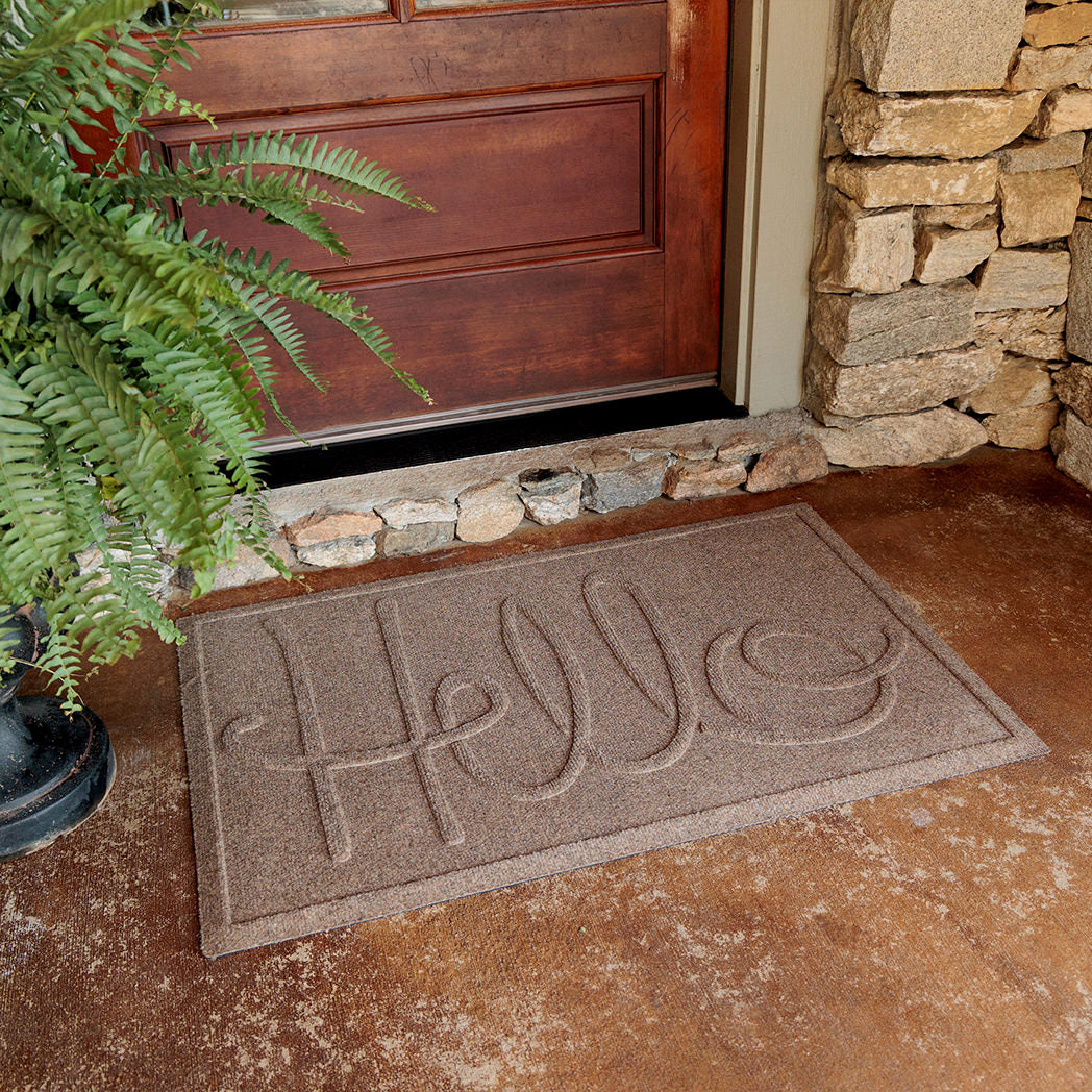 The WaterHog Hello Doormat sits outside a wooden front door, surrounded by stone siding and a potted fern. The mat is a tan color with the word “Hello” written in large, looping raised letters across its surface. Its textured design and sturdy build stand out against the rough concrete porch, giving a warm and inviting first impression.