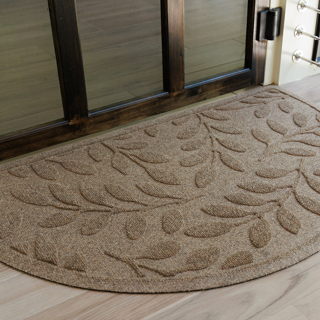 Close up of semi-circle WaterHog doormat with a bi-level leaf pattern on a wooden floor in front of a glass door.