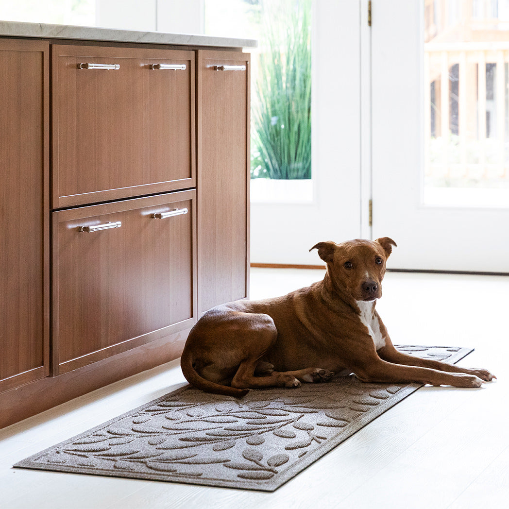 Dog laying on a leafy-textured runner in a kitchen with wooden cabinets