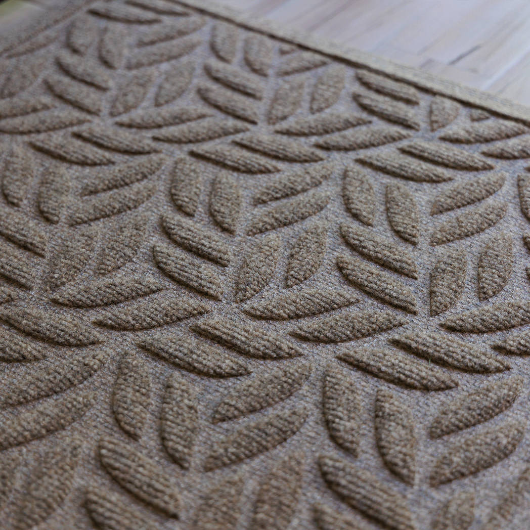 The WaterHog Canopy Doormat in a warm camel tone features a repeating leaf-inspired pattern, with overlapping curved shapes that create the look of a gentle canopy of foliage. The raised texture adds depth and dimension, giving it both a natural, organic feel and durable functionality for trapping dirt and moisture.