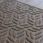 The WaterHog Canopy Doormat in a warm camel tone features a repeating leaf-inspired pattern, with overlapping curved shapes that create the look of a gentle canopy of foliage. The raised texture adds depth and dimension, giving it both a natural, organic feel and durable functionality for trapping dirt and moisture.