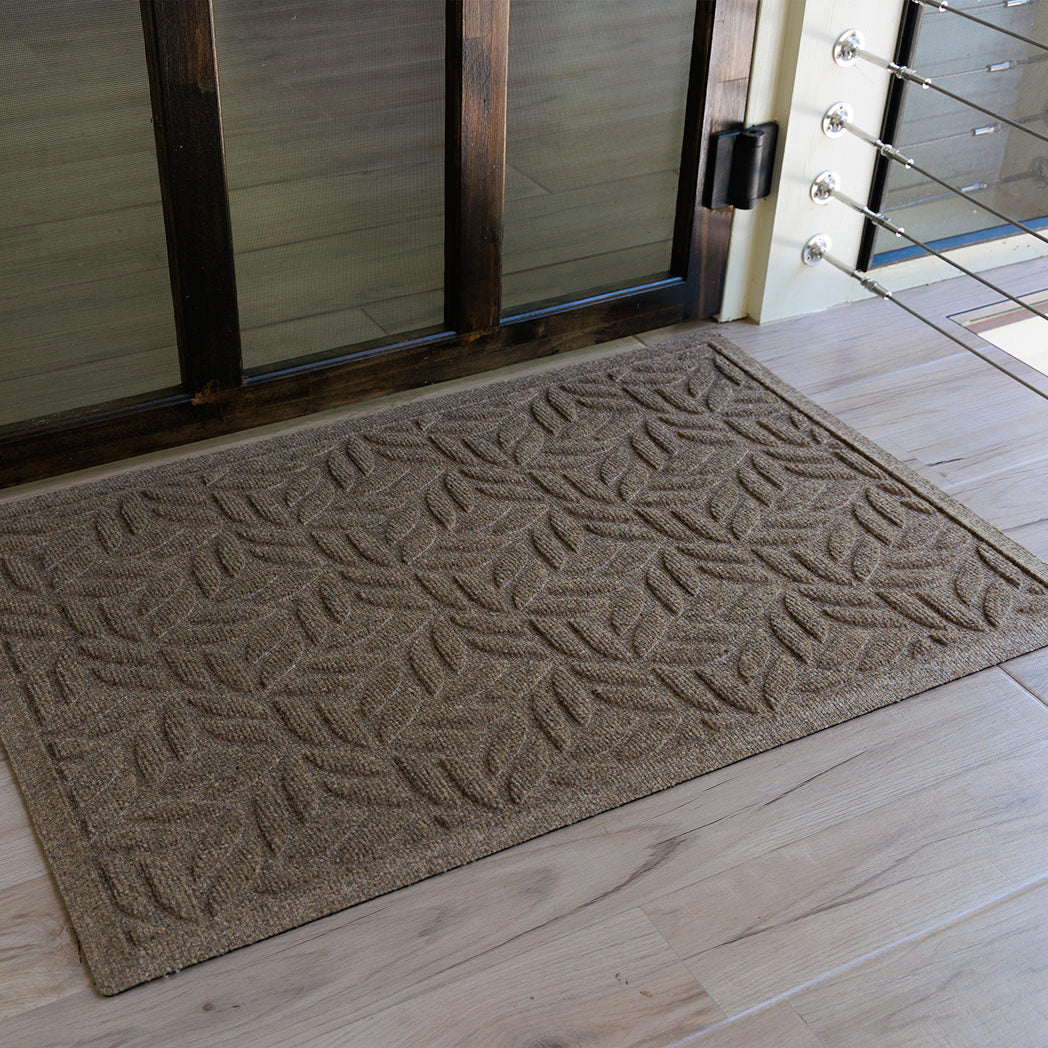 The WaterHog Canopy Doormat in a warm tan shade rests inside a doorway with light wood flooring. Its raised leaf-like pattern forms a repeating, organic design that resembles overlapping branches or fronds, combining natural elegance with the mat’s signature durability and water-trapping texture.