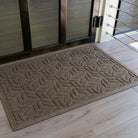 The WaterHog Canopy Doormat in a warm tan shade rests inside a doorway with light wood flooring. Its raised leaf-like pattern forms a repeating, organic design that resembles overlapping branches or fronds, combining natural elegance with the mat’s signature durability and water-trapping texture.