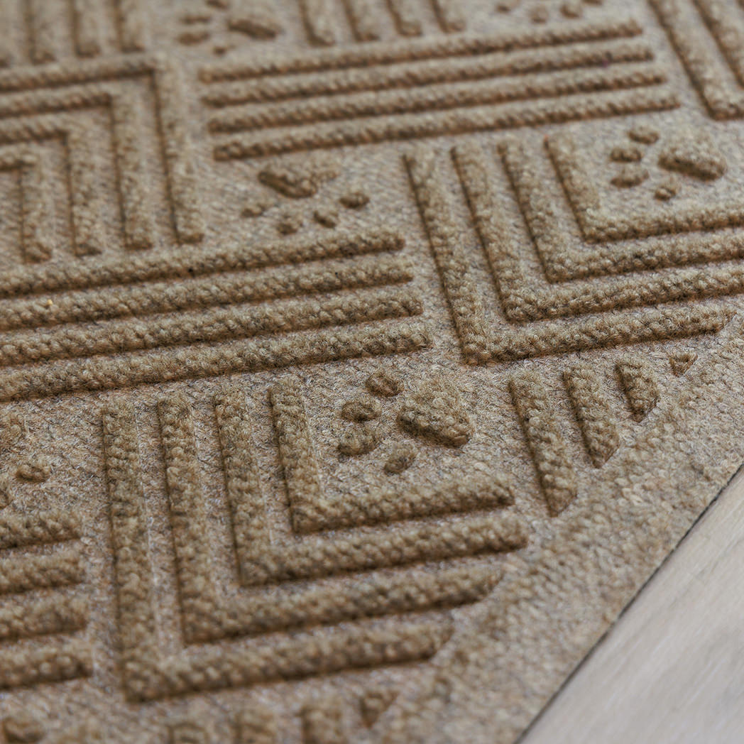 Angled close-up of a tan WaterHog Cat Litter Mat on light flooring, showing its raised edge and detailed surface pattern of paw prints and intersecting ridges.