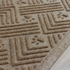 Angled close-up of a tan WaterHog Cat Litter Mat on light flooring, showing its raised edge and detailed surface pattern of paw prints and intersecting ridges.
