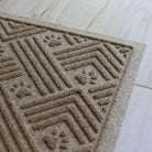 Close-up of the corner of a tan WaterHog Cat Litter Mat on light wood flooring, showing the raised edge and textured surface with paw print and line patterns.