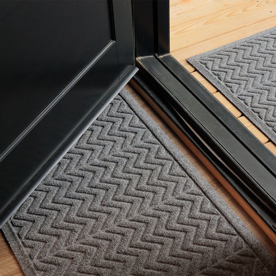 The WaterHog Chevron Doormat and Runner in a cool gray tone sit on either side of a black-framed door. The mats feature a raised zigzag pattern that repeats across the surface, creating a sharp, modern look. The texture is slightly ridged to help catch dirt, while the gray color pairs seamlessly with the light wood flooring and dark door frame for a balanced, clean entryway style.