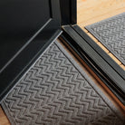 The WaterHog Chevron Doormat and Runner in a cool gray tone sit on either side of a black-framed door. The mats feature a raised zigzag pattern that repeats across the surface, creating a sharp, modern look. The texture is slightly ridged to help catch dirt, while the gray color pairs seamlessly with the light wood flooring and dark door frame for a balanced, clean entryway style.