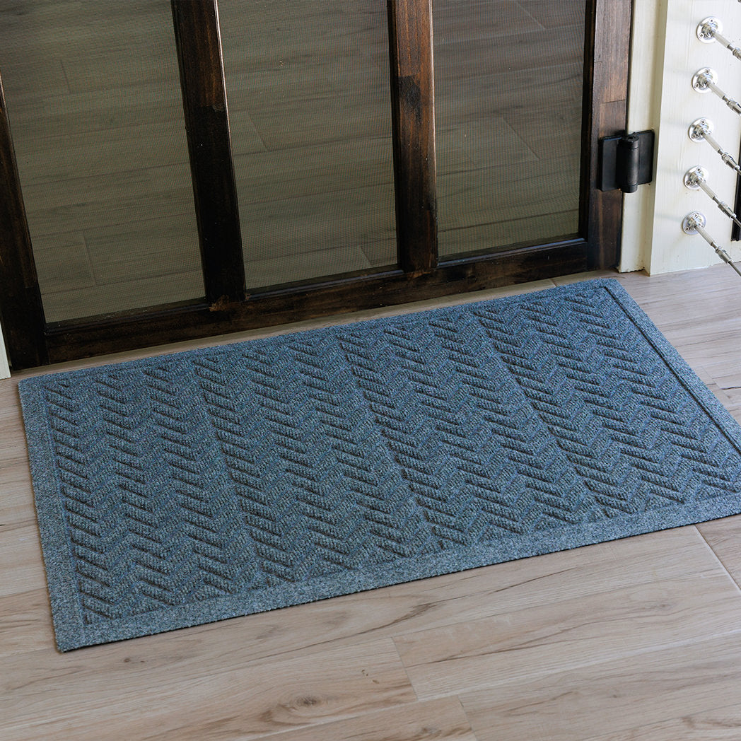 The WaterHog Chevron Doormat in a blue-grey tone sits at the base of a wooden-framed glass door. Its repeating V-shaped pattern runs in neat horizontal rows, creating a crisp, textured surface that contrasts softly with the pale wood floor, offering both modern style and practical dirt-trapping design.