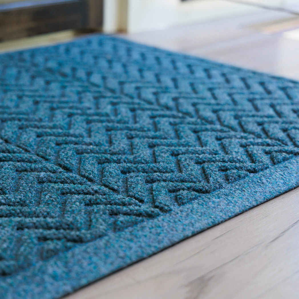 A close-up of the WaterHog Chevron Doormat in deep blue shows its tightly woven zigzag ridges and low-profile border. The textured fibers catch the light, highlighting the mat’s raised V-shaped pattern designed to trap dirt and moisture while maintaining a sleek, structured look against the light wood flooring.