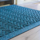 A close-up of the WaterHog Chevron Doormat in deep blue shows its tightly woven zigzag ridges and low-profile border. The textured fibers catch the light, highlighting the mat’s raised V-shaped pattern designed to trap dirt and moisture while maintaining a sleek, structured look against the light wood flooring.