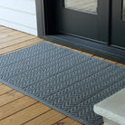 The WaterHog Chevron Doormat in gray sits outside a black-framed glass door on a wooden deck. The mat’s surface features a repeating V-shaped zigzag pattern that runs neatly in rows, creating a modern, textured look. The cool gray tone stands out against the warm wood planks, adding a clean, structured touch to the outdoor entryway.