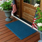 The WaterHog Chevron Doormat in a rich navy blue color sits outside a wooden front door on a red-stained porch. The mat’s zigzag pattern adds subtle movement and texture beneath two potted plants and small American flags, creating a welcoming, patriotic entryway that blends classic charm with durable function.