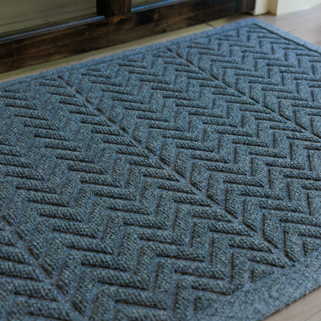 The WaterHog Chevron Doormat in a blue-grey hue rests on light wood flooring by a dark-framed glass door. Its raised zigzag pattern catches the light, adding depth and contrast to the cool-toned fibers, creating a clean and modern look that combines functionality with subtle texture.