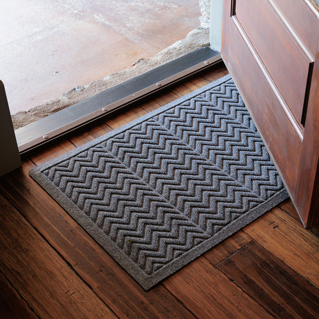 The WaterHog Chevron Doormat in gray rests at the base of a wooden door. Its raised zigzag pattern runs across the mat, creating a bold, rhythmic texture designed to trap dirt and water. The soft gray tone complements the rustic wood floor and stone threshold, giving the entryway a clean, balanced look that blends function with subtle style.