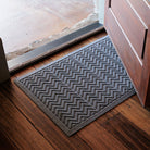 The WaterHog Chevron Doormat in gray rests at the base of a wooden door. Its raised zigzag pattern runs across the mat, creating a bold, rhythmic texture designed to trap dirt and water. The soft gray tone complements the rustic wood floor and stone threshold, giving the entryway a clean, balanced look that blends function with subtle style.