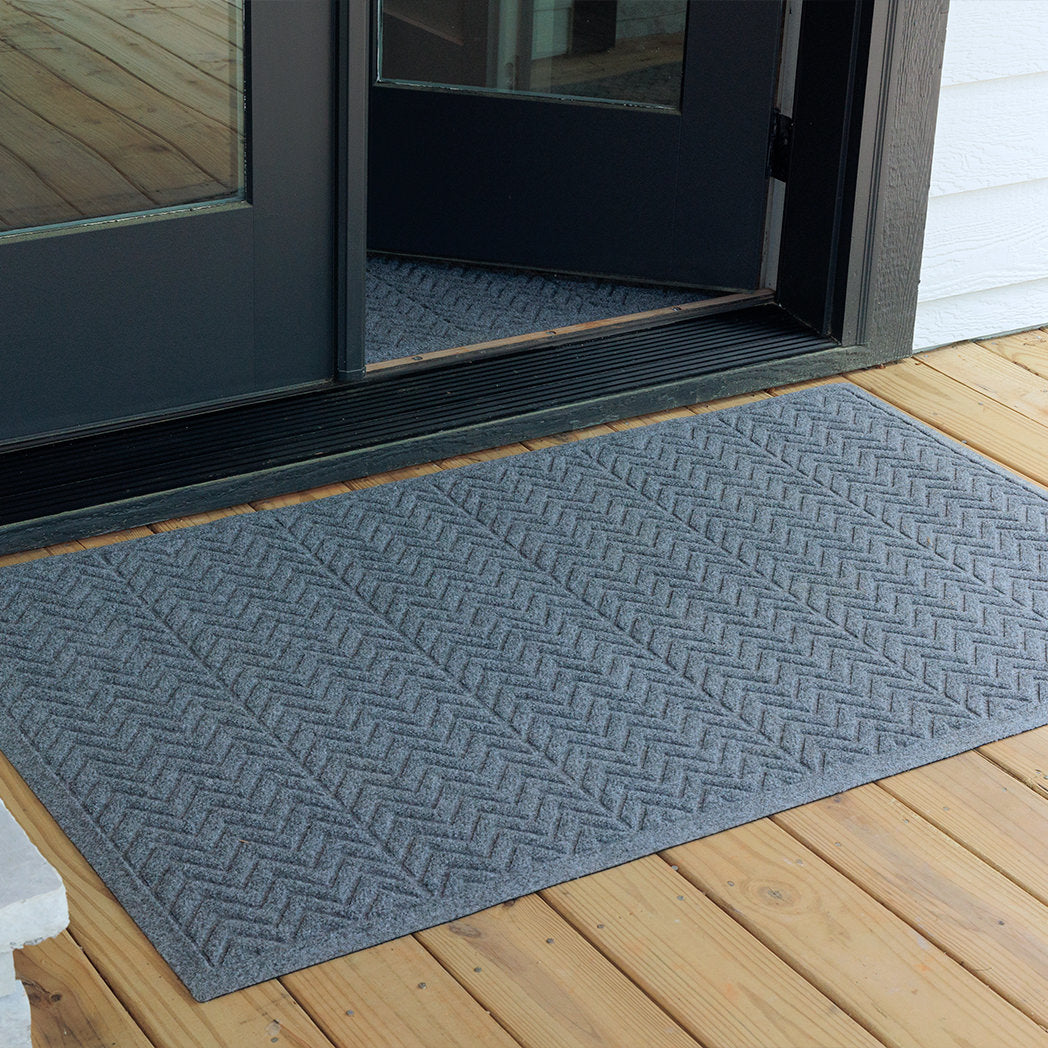 The WaterHog Chevron Doormat in soft blue gray sits neatly outside a black-framed glass door on a wooden deck. Its surface features a clean zigzag pattern arranged in wide horizontal panels, giving it a structured, modern look that blends durability with subtle texture for a welcoming outdoor entryway.