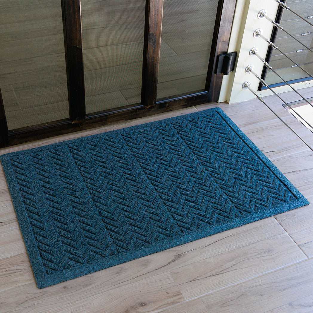 The WaterHog Chevron Doormat in a deep blue color rests on light wood flooring near a glass door. The surface features a repeating V-shaped zigzag pattern that gives the mat texture and grip. The rich blue hue adds a cool, modern accent to the entryway, contrasting softly with the warm-toned floor and dark wood trim of the door.