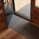 The WaterHog Chevron Runner and Doormat in gray sit at a doorway with one mat indoors and one just outside. Their raised zigzag pattern forms a continuous chevron texture that adds movement and dimension. The soft gray color contrasts the dark door and warm wood flooring, giving the entryway a polished and cohesive look while keeping dirt and moisture contained.