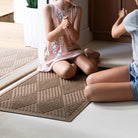 The WaterHog Diamonds Doormat in a camel tan color sits beneath two children playing near a doorway. Its raised diamond pattern forms a repeating grid of diagonally lined shapes that add depth and texture to the mat’s surface. The warm neutral tone pairs naturally with the light flooring, creating a clean, inviting entryway that feels both soft and structured